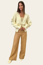 Load image into Gallery viewer, Woman in a cream cardigan and beige pants standing in a room with a mirror and plants.